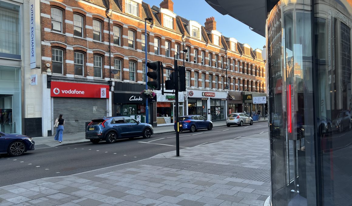 Street view of a row of red brick shops (Vodafone, Chipotle, Clarks, McDonald's) under a blue sky with cars parked along the curb.