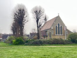 Brick church with Gothic arched windows and a cross on the roof, framed by bare trees over a green lawn under an overcast sky.