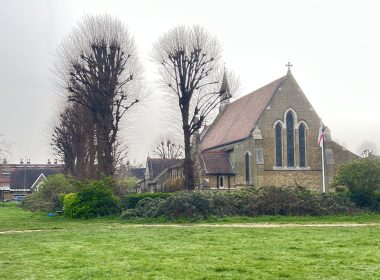 Brick church with Gothic arched windows and a cross on the roof, framed by bare trees over a green lawn under an overcast sky.