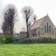 Brick church with Gothic arched windows and a cross on the roof, framed by bare trees over a green lawn under an overcast sky.
