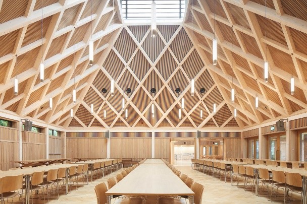 Interior of a large wooden hall with a crisscross beam roof, long tables, and rows of chairs along the sides under a skylight.