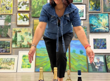 Smiling woman in a denim top bends toward four wine bottles arranged on the gallery floor, with colorful paintings on the wall behind her.
