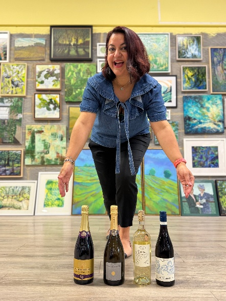 Smiling woman in a denim top bends toward four wine bottles arranged on the gallery floor, with colorful paintings on the wall behind her.