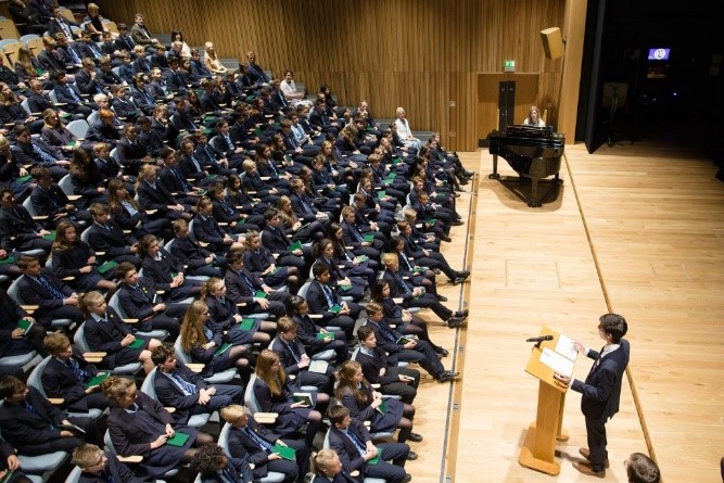 Large lecture hall filled with hundreds of suited attendees facing a speaker at a wooden podium on stage; a pianist sits at a grand piano nearby.