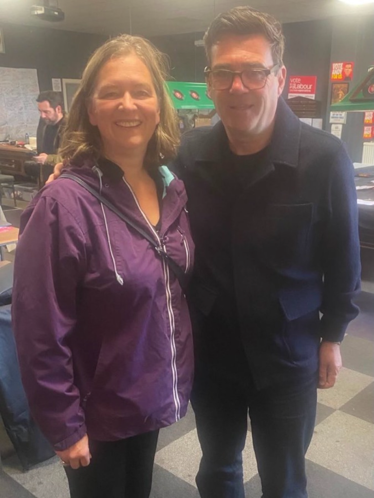 Two adults posing together indoors, smiling, with posters and office furniture in the background. The woman wears a purple jacket; the man wears a dark coat and glasses.