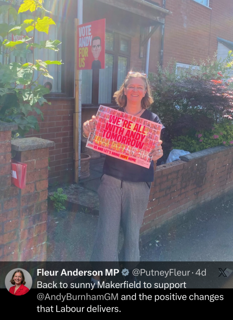 Woman standing outside a brick house, holding a political poster that reads 'WE'RE ALL WITH ANDY TOMORROW'.