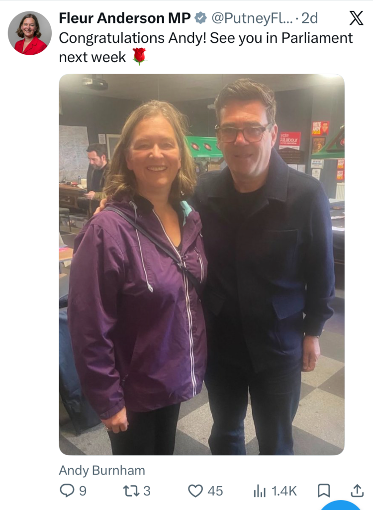 Smiling woman in a purple jacket standing next to a man in a dark coat indoors, with posters and a map visible in the background, posing for a selfie.