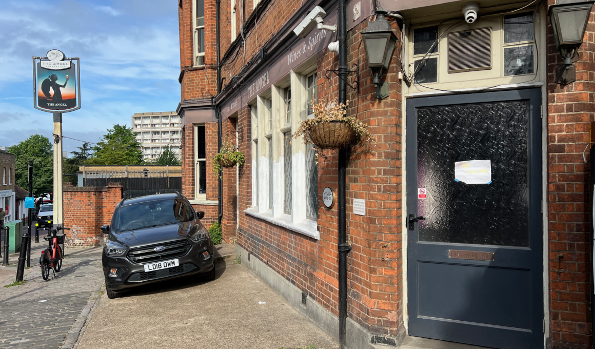 Brick pub exterior with a dark blue door labeled 'Public Bar', hanging plant basket, and a black SUV parked outside on a sunny street.