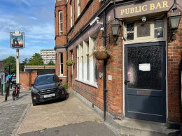 Brick pub exterior with a dark blue door labeled 'Public Bar', hanging plant basket, and a black SUV parked outside on a sunny street.