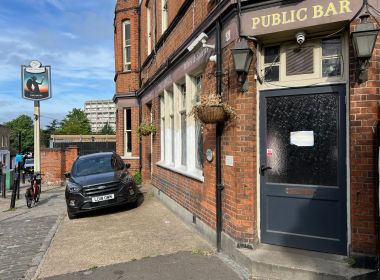 Brick pub exterior with a dark blue door labeled 'Public Bar', hanging plant basket, and a black SUV parked outside on a sunny street.