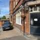 Brick pub exterior with a dark blue door labeled 'Public Bar', hanging plant basket, and a black SUV parked outside on a sunny street.