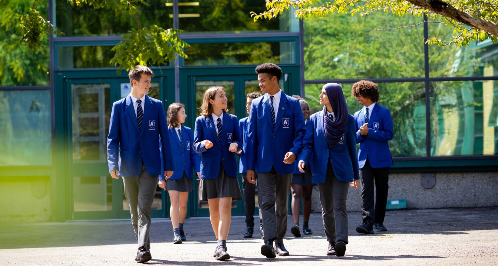 Group of students in blue uniforms walking together outside a glass-front school building on a sunny day.