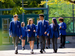 Group of students in blue uniforms walking together outside a glass-front school building on a sunny day.