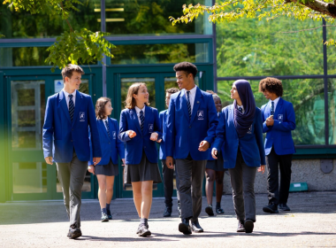 Group of students in blue uniforms walking together outside a glass-front school building on a sunny day.