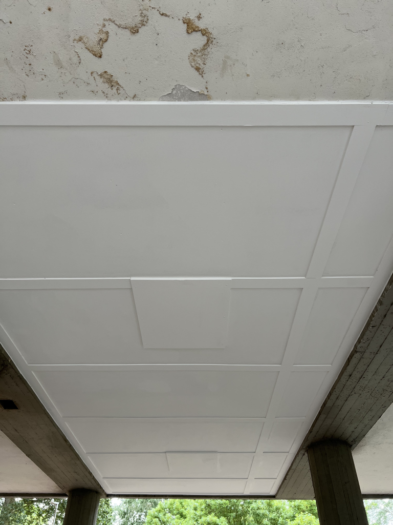 White coffered ceiling panels along an outdoor covered walkway, with concrete columns and greenery visible outside