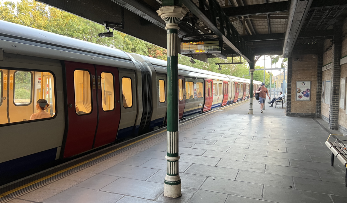 Train at a platform with a row of doors open to a warm-lit carriage; a green and white pillar stands in foreground under a metal canopy.