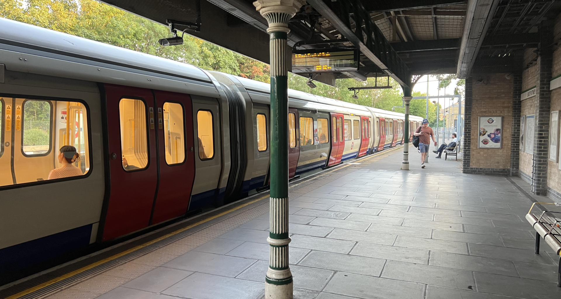Train at a platform with a row of doors open to a warm-lit carriage; a green and white pillar stands in foreground under a metal canopy.