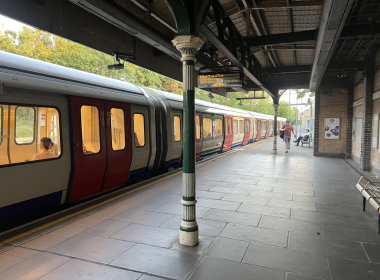 Train at a platform with a row of doors open to a warm-lit carriage; a green and white pillar stands in foreground under a metal canopy.