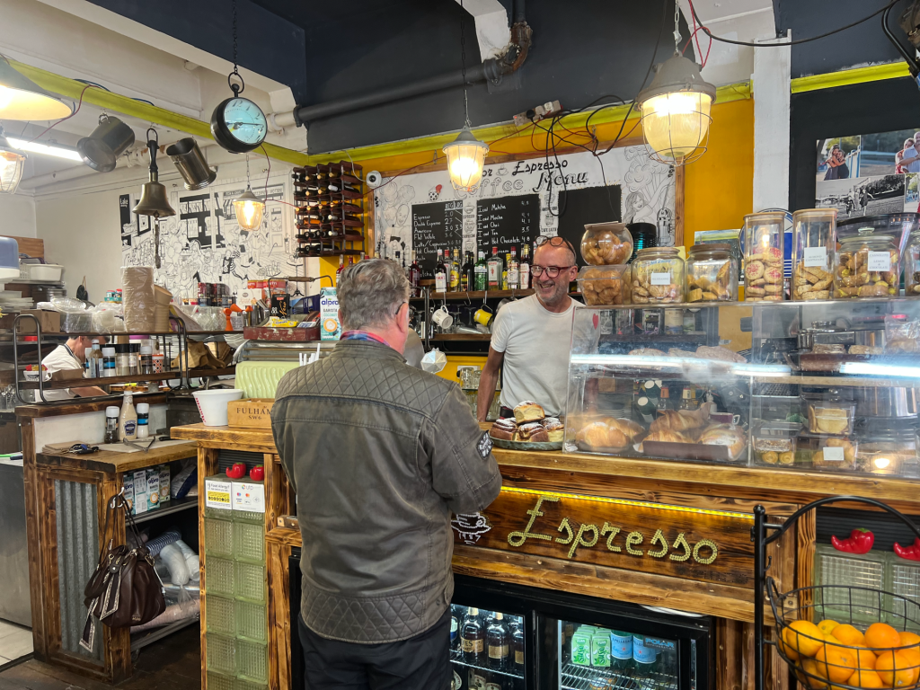 Café scene with a staff member behind a pastry counter, glass display of baked goods and jars, chalkboard menu in the background.