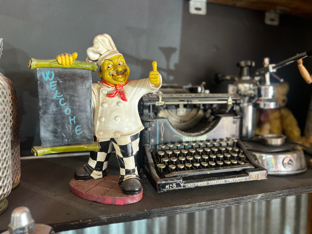 Whimsical chef statue giving a thumbs-up, leaning on a chalky block beside an antique Underwood typewriter on a shelf.
