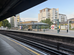 Platform at East Putney station with tracks in the foreground and a white mid-rise building across the street against a blue sky under a dark overpass the left side.