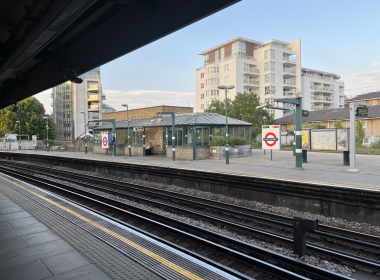 Platform at East Putney station with tracks in the foreground and a white mid-rise building across the street against a blue sky under a dark overpass the left side.