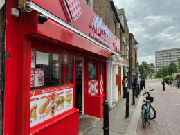 Bright red fast-food restaurant storefront with menu posters in the window and people walking along the sidewalk outside.