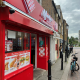 Bright red fast-food restaurant storefront with menu posters in the window and people walking along the sidewalk outside.