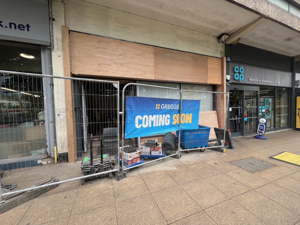 Storefront under renovation with plywood boards, construction fencing, and a 'COMING SOON' banner in front of a Co-op shop.