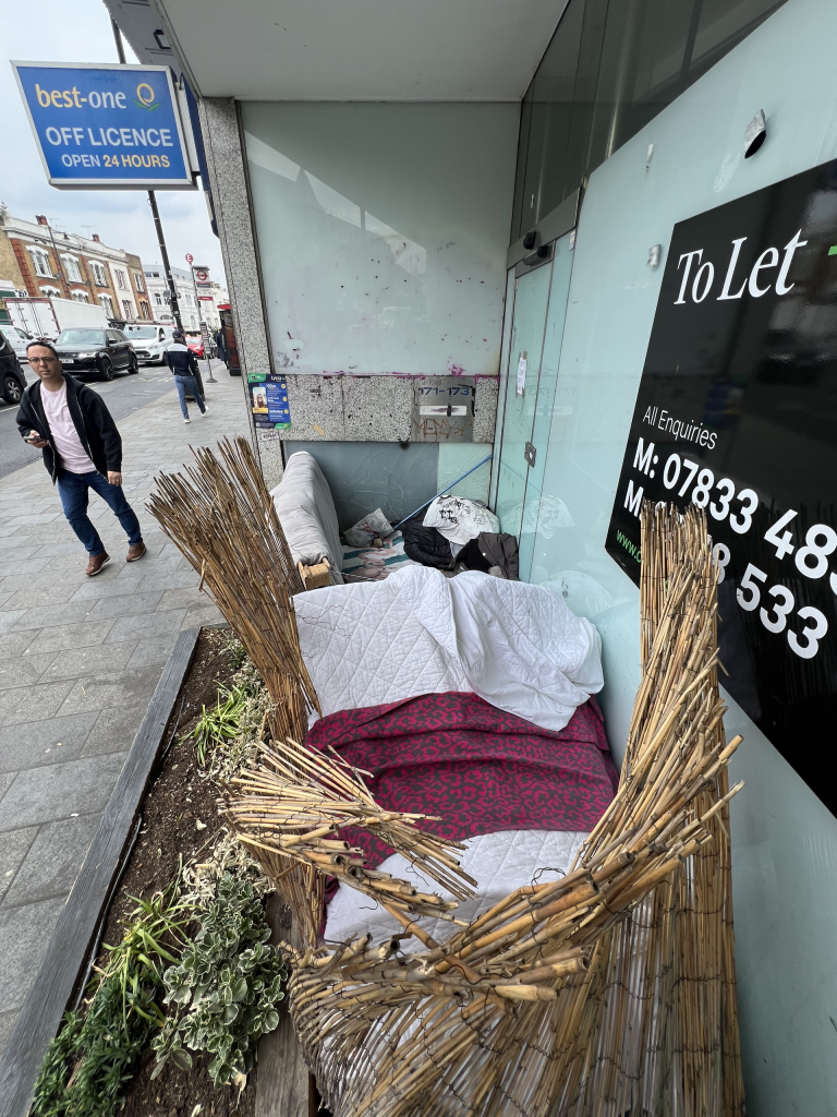 Storefront with a 'To Let' sign; in front, wicker barrier and cushions/blankets create a makeshift seating area beside a planter along the shopfront, pedestrians and street scene in background.