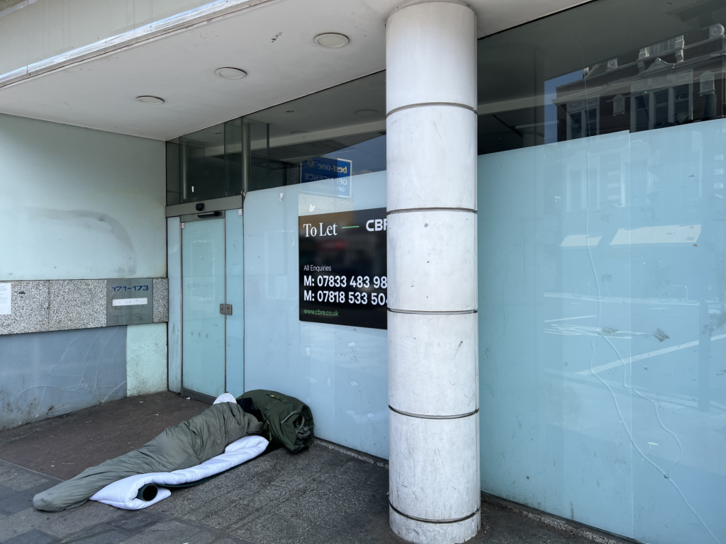 Person sleeping on a blanket on the sidewalk in front of a glass storefront beside a large pillar.