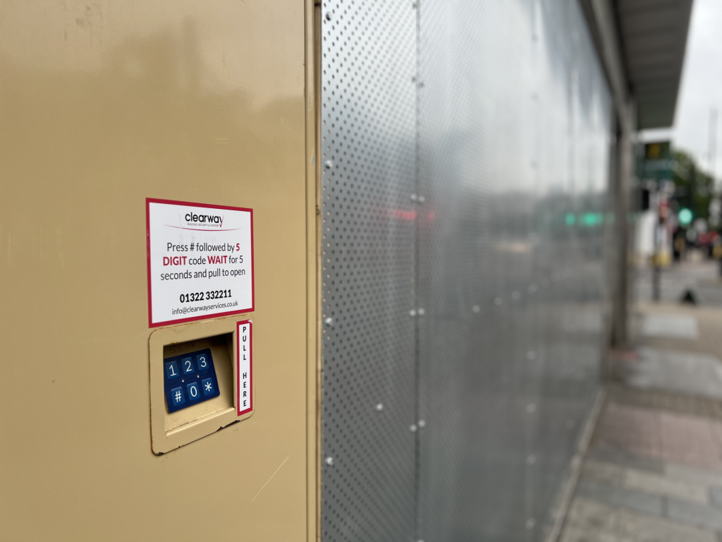 Beige wall-mounted keypad with a blue numeric keypad and a 'CLEARWAY' sticker describing press-and-wait instructions next to a perforated metal panel.