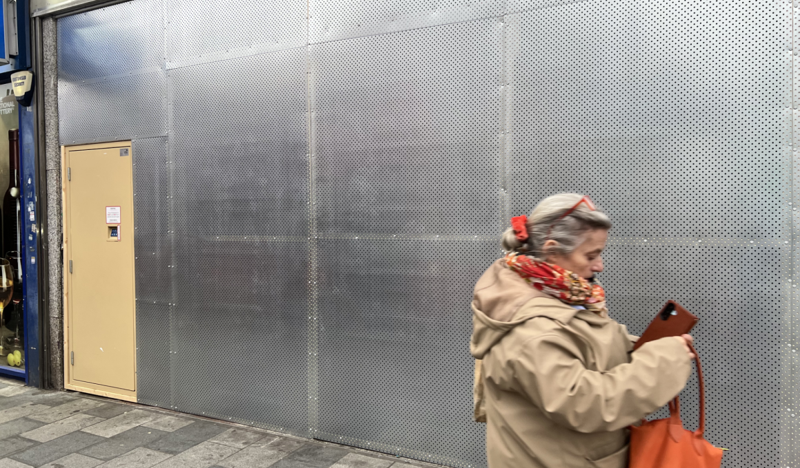 Urban storefront with a perforated metal wall; beige door on the left and a passerby in a beige coat carrying an orange bag in the foreground