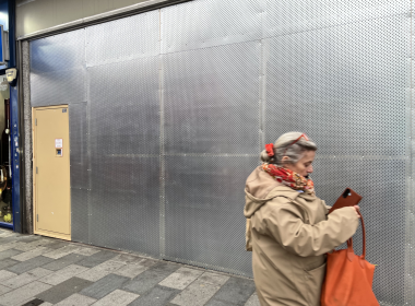 Urban storefront with a perforated metal wall; beige door on the left and a passerby in a beige coat carrying an orange bag in the foreground
