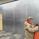 Urban storefront with a perforated metal wall; beige door on the left and a passerby in a beige coat carrying an orange bag in the foreground