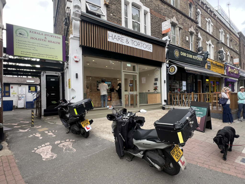 Row of shops on a busy street with Hare & Tortoise, Kerala Ayurveda, and Dim Sum Kitchen signs, and two motorbikes parked at the curb.