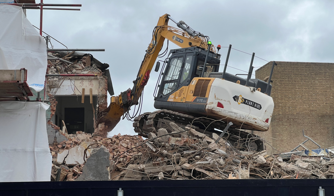 Yellow excavator atop a rubble pile at a building demolition site, with a partially collapsed brick wall and scaffolding nearby.