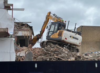 Yellow excavator atop a rubble pile at a building demolition site, with a partially collapsed brick wall and scaffolding nearby.