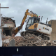 Yellow excavator atop a rubble pile at a building demolition site, with a partially collapsed brick wall and scaffolding nearby.