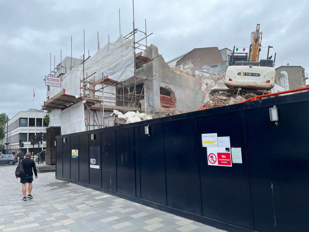 Construction site behind a tall black barrier with scaffolding and white tarps, and a yellow excavator on rubble.