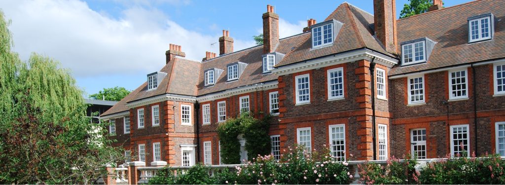 Red brick townhouse with white-framed windows, ivy over the entry, and rose bushes along a low fence beneath a blue sky.