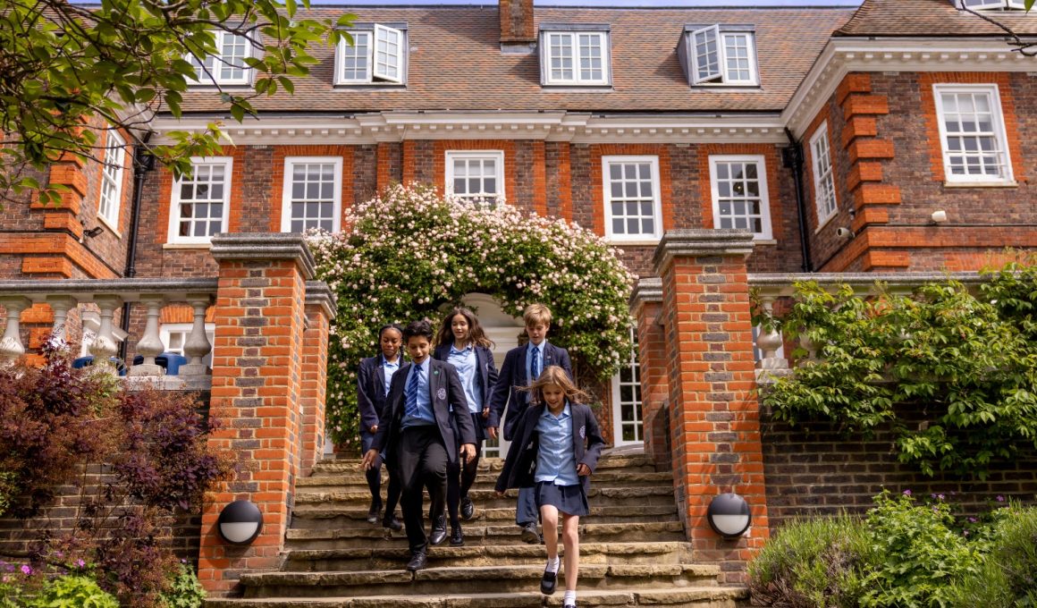 Group of students in navy blazers and blue shirts walking down stone steps from a red-brick school, under a flowering arch.