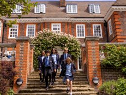 Group of students in navy blazers and blue shirts walking down stone steps from a red-brick school, under a flowering arch.