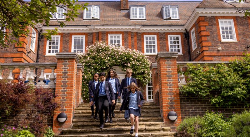 Group of students in navy blazers and blue shirts walking down stone steps from a red-brick school, under a flowering arch.