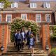 Group of students in navy blazers and blue shirts walking down stone steps from a red-brick school, under a flowering arch.