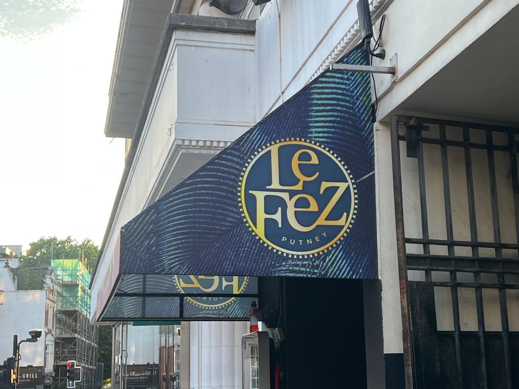 Storefront banner with a blue circular logo reading 'Le Dez' (Putney) on a blue patterned awning, next to a metal gate.