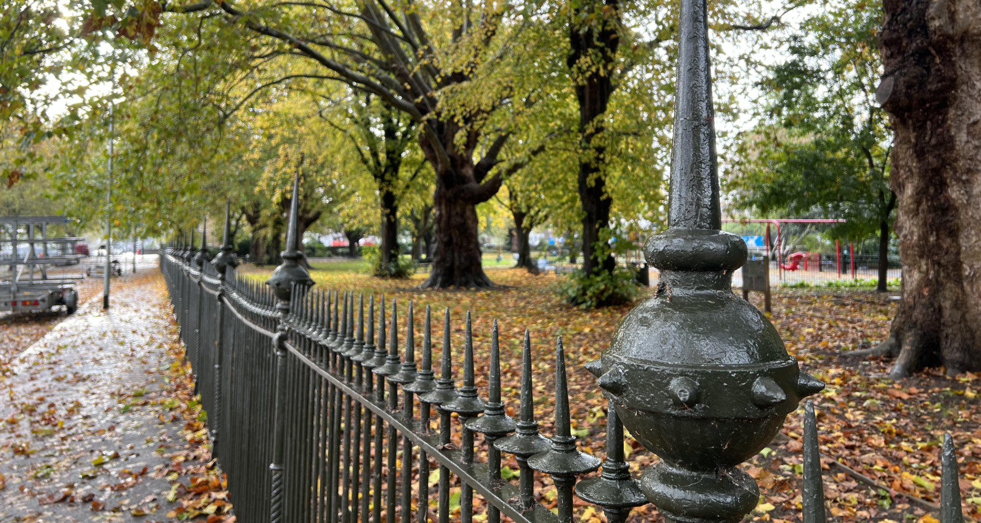 Wrought-iron fence along a leaf-covered sidewalk in a park with autumn trees in the background.