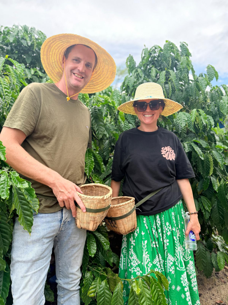 Man and woman in straw hats smiling among coffee plants, holding woven baskets in a lush field.