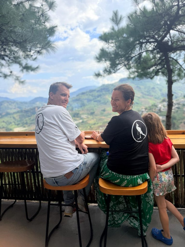 Family of three sitting at a wooden railing, smiling at the camera with a mountain valley in the background.