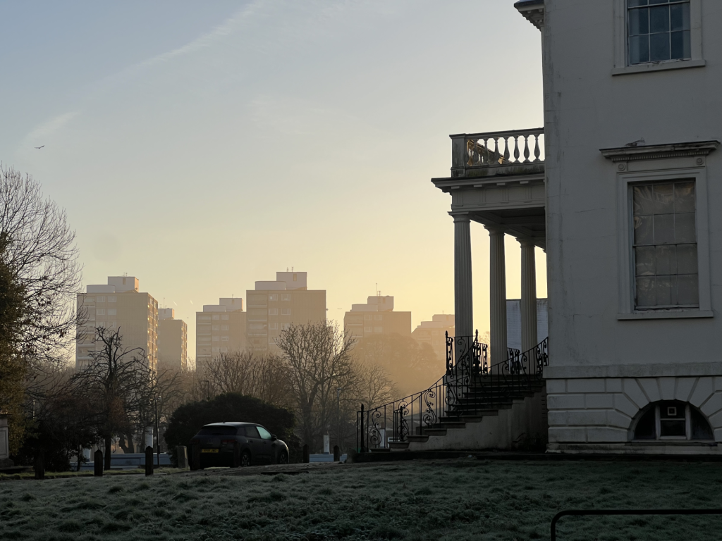 Sunset view of a grand white building with columns and a balcony on the right, overlooking a cityscape with bare trees and a parked car.